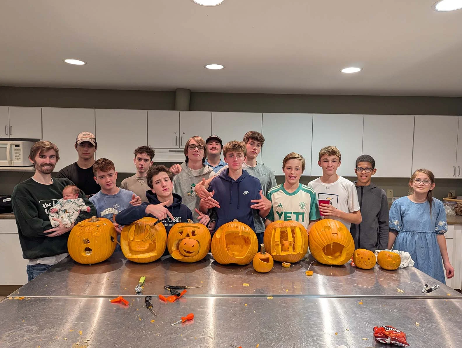 A group of about 13 boys with 2 adults standing in a large kitchen with their carved out pumpkins. They're trying to look cool.