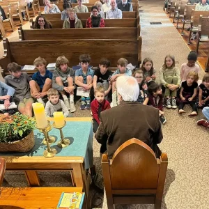 A man reads to children of all ages up front in the church during the children's sermon.